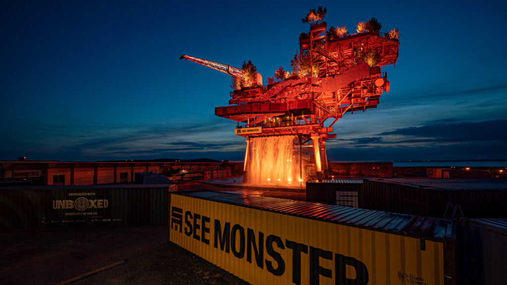 A giant oil rig covered in a man-made garden with plants and waterfalls, built high on top of shipping containers on the shore of Weston-Super-Mare with signage showing an example of marketing big culture events